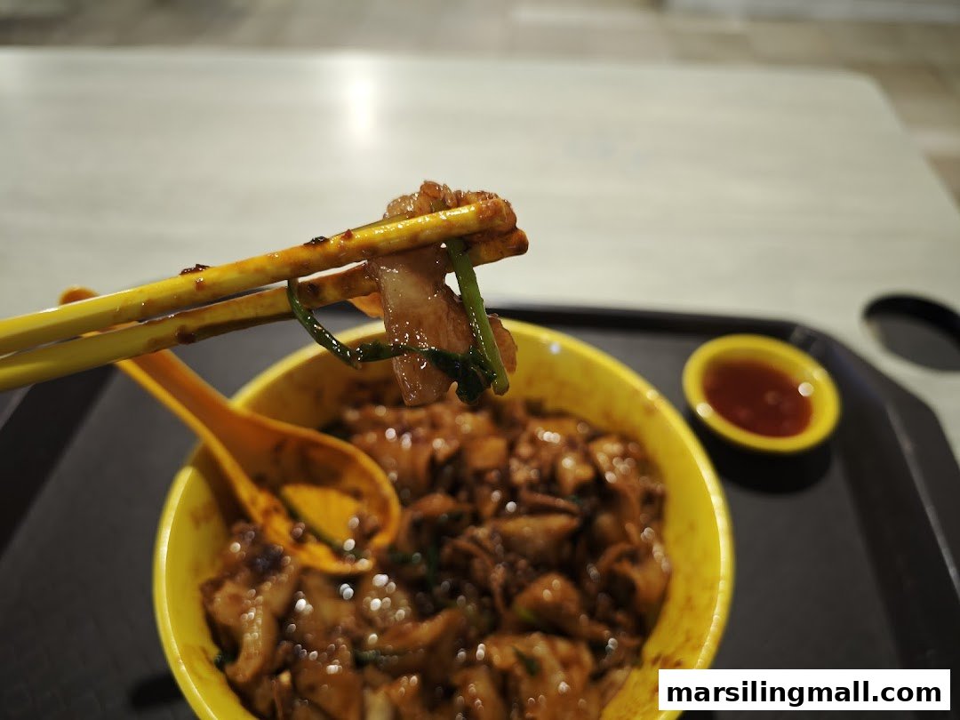Chopsticks lifting a piece of stir-fried kway teow from a yellow bowl, with a side of chili sauce.