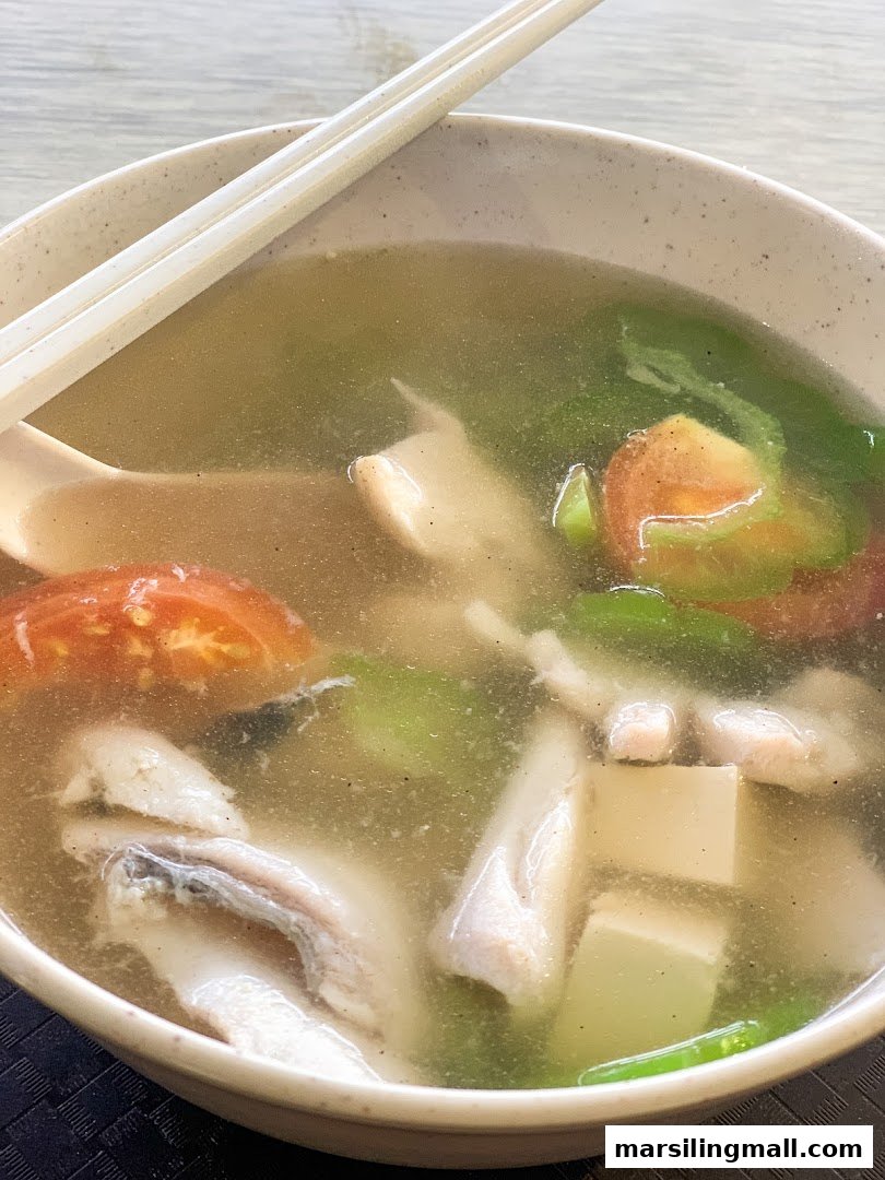 A close-up of a bowl of clear fish soup with fish slices, tomatoes, tofu, and green vegetables.