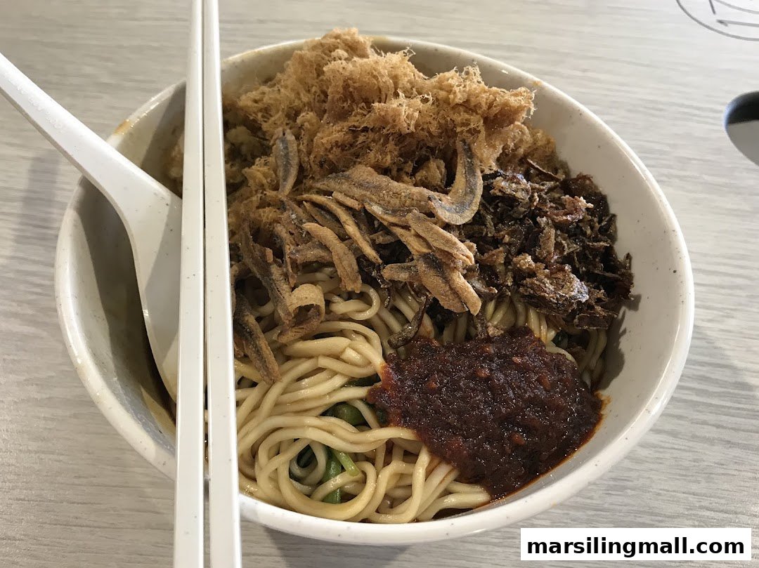 A close-up of a bowl of noodles topped with crispy anchovies, fried shallots, and chili paste.