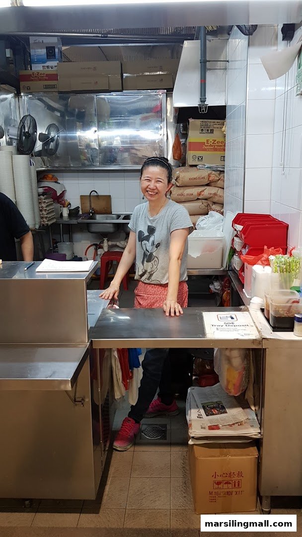 A smiling staff member stands behind a counter in a clean, bustling kitchen area.