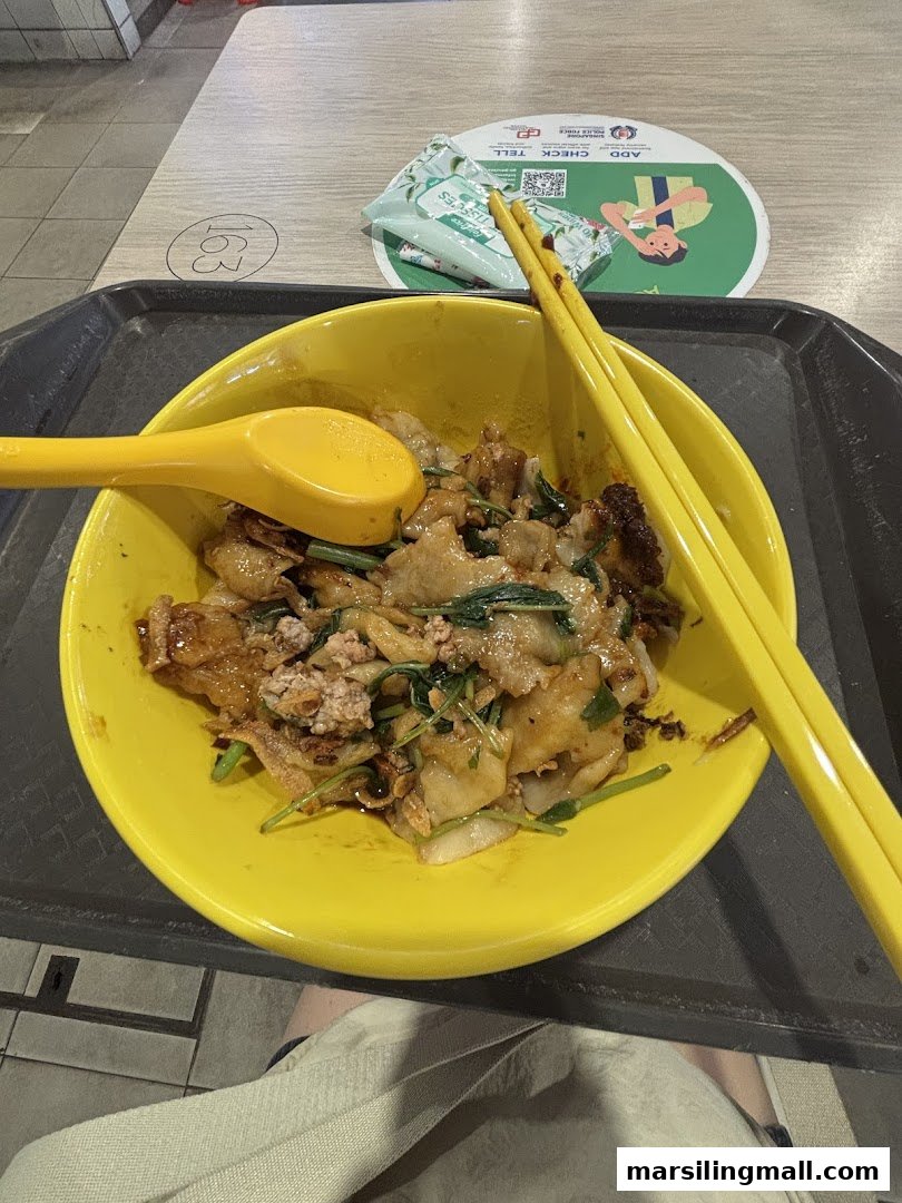 A vibrant yellow bowl filled with stir-fried noodles, minced meat, and green vegetables, served with chopsticks and a spoon.