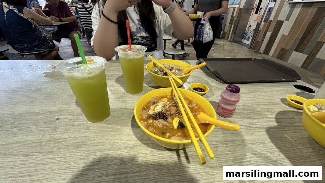 Two bowls of noodle soup and two green drinks on a light wooden table inside a bustling eatery.