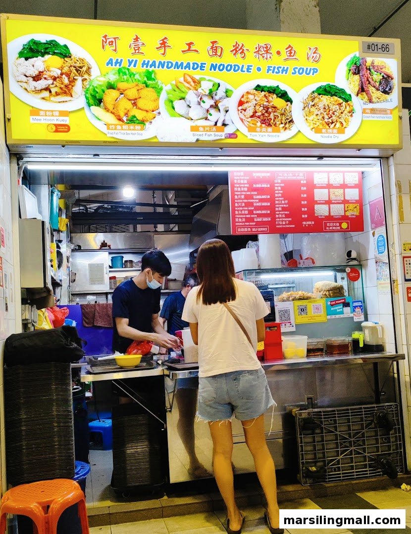 A vibrant hawker stall, 'Ah Yi Handmade Noodle Fish Soup,' with staff serving customers and a prominent signboard.