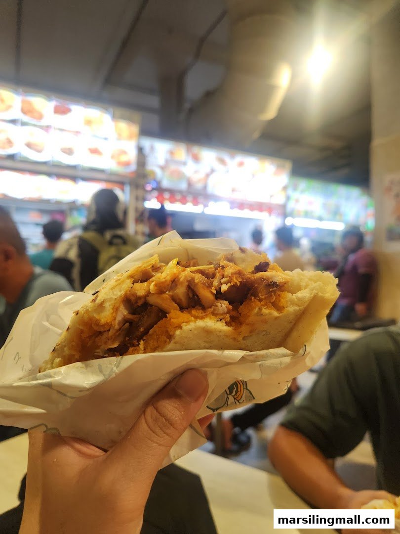 A hand holds a delicious, freshly prepared kebab in a bustling food court setting.