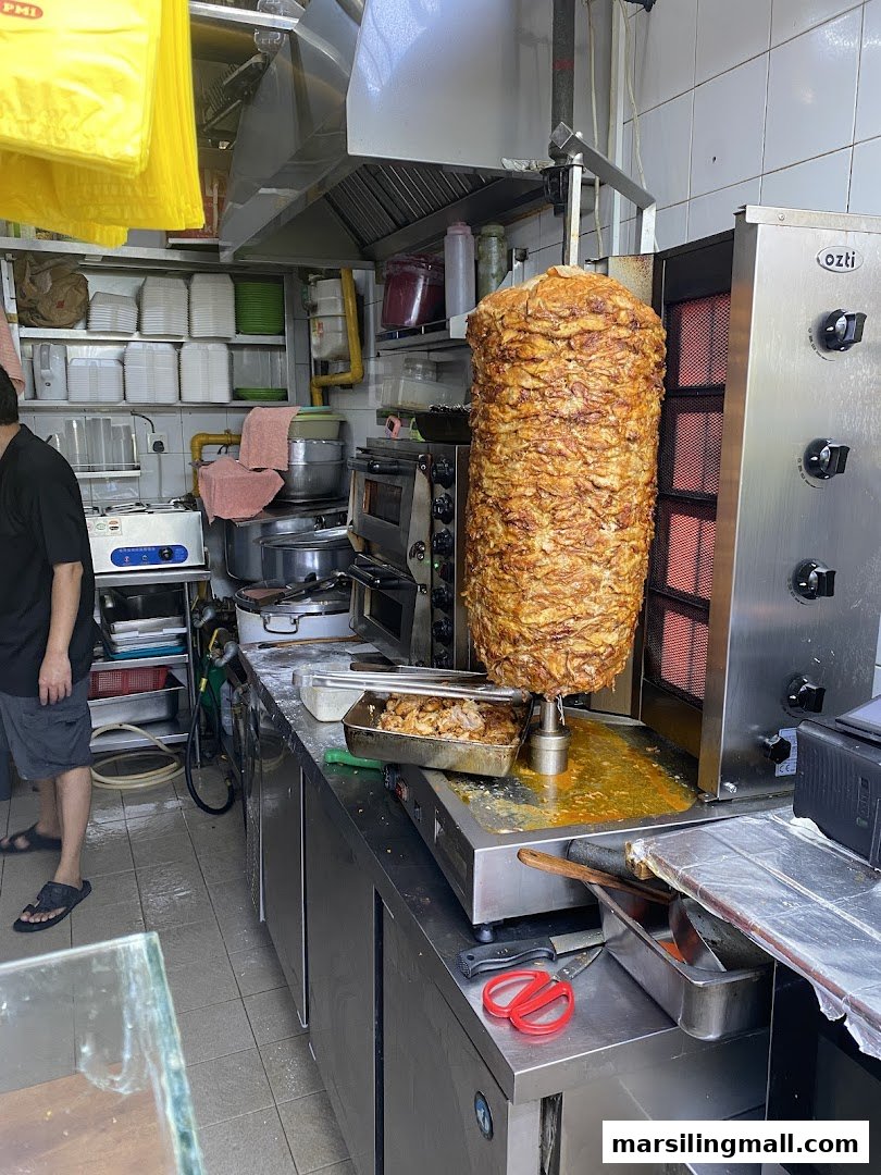 A large rotating kebab spit with meat cooking, inside a bustling kitchen area.