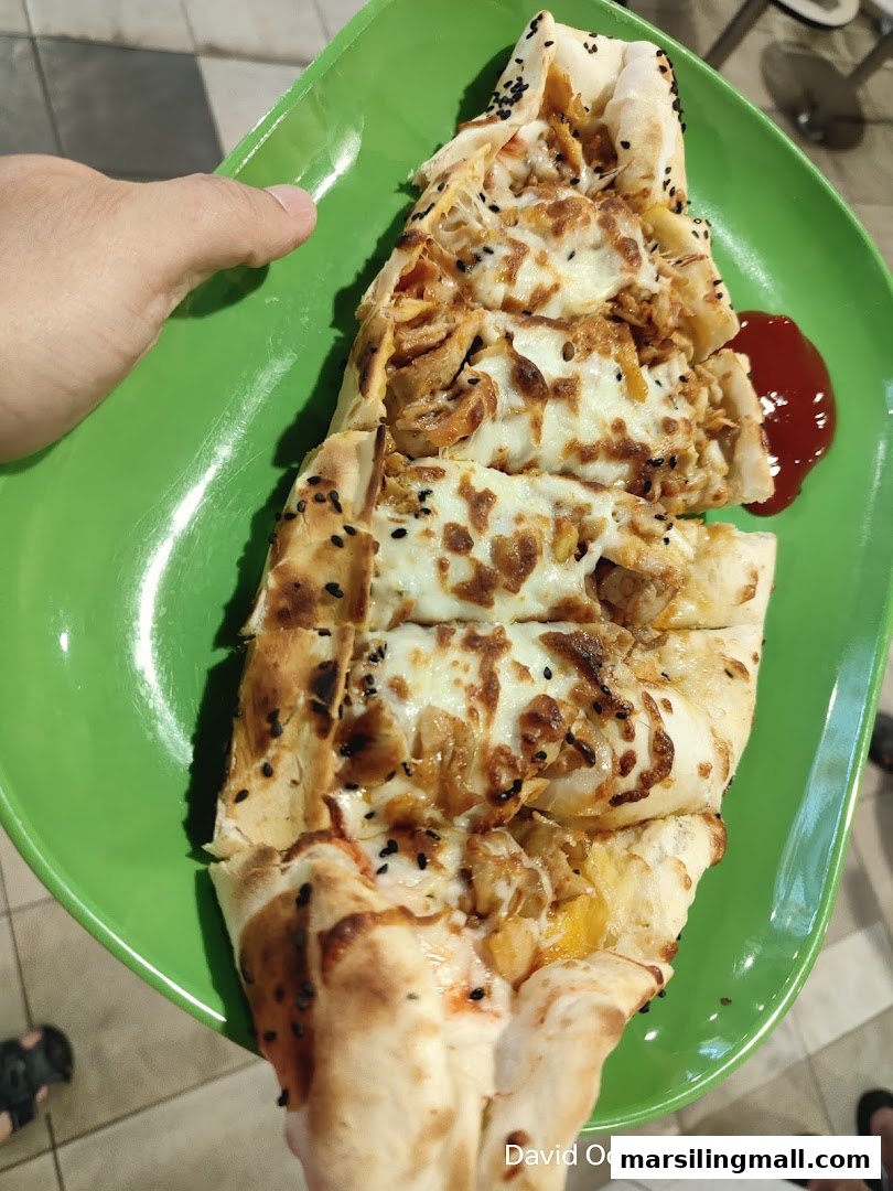 A delicious boat-shaped flatbread topped with chicken, melted cheese, and black seeds, served on a green plate.
