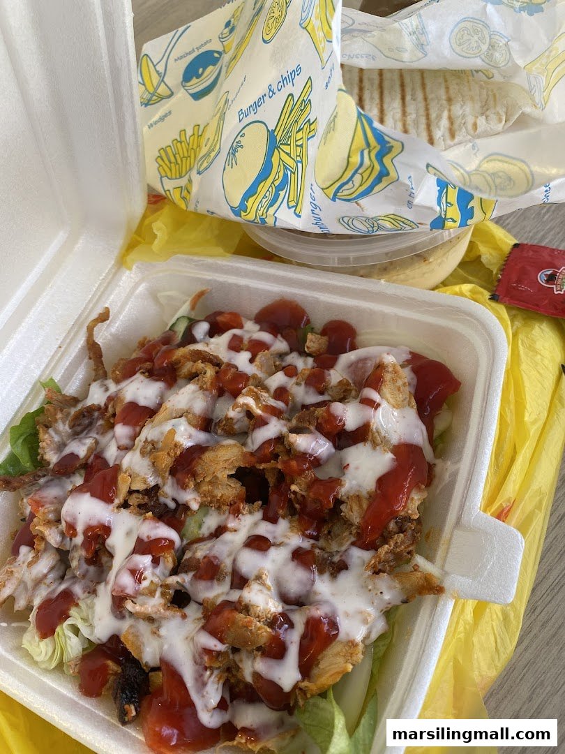 A takeaway container filled with chicken kebab meat, lettuce, ketchup, and white sauce, with a burger wrapper in the background.