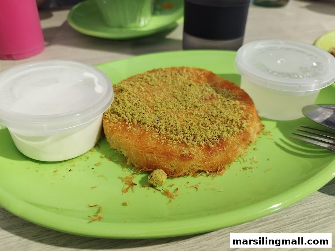 A delicious kunafa dessert topped with pistachios, served with cream and syrup on a vibrant green plate.