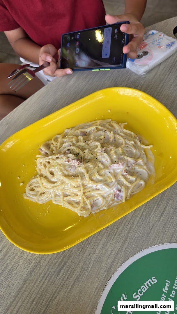 A creamy pasta dish served in a bright yellow plate, with a person using a phone nearby.