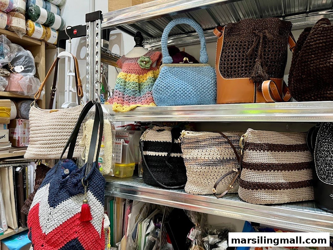 A shelf displaying various handcrafted crochet bags, a small dress, and yarn in a crafting shop.