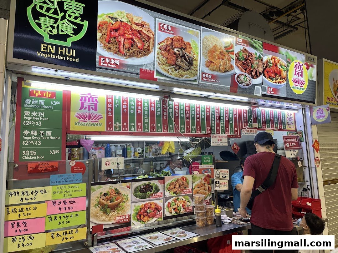 A customer orders vegetarian food from a brightly lit stall displaying various dishes and prices.