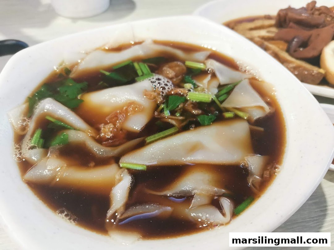 A close-up of a bowl of kway chap with broad rice noodles in dark broth, garnished with herbs.