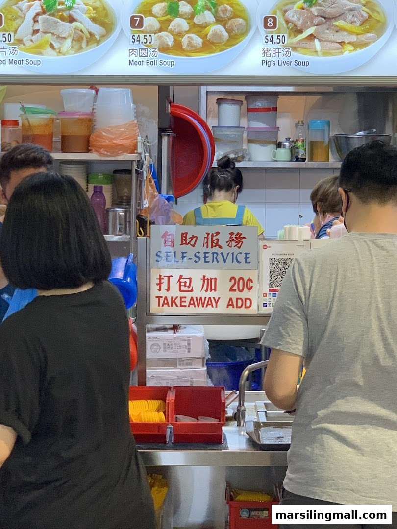 A bustling food stall counter with customers and a clear menu board displaying soup options.