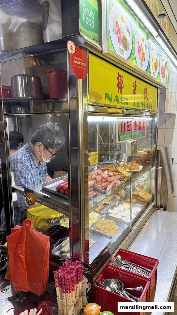 A bustling food stall counter showcasing various Nasi Lemak ingredients and a staff member preparing orders.