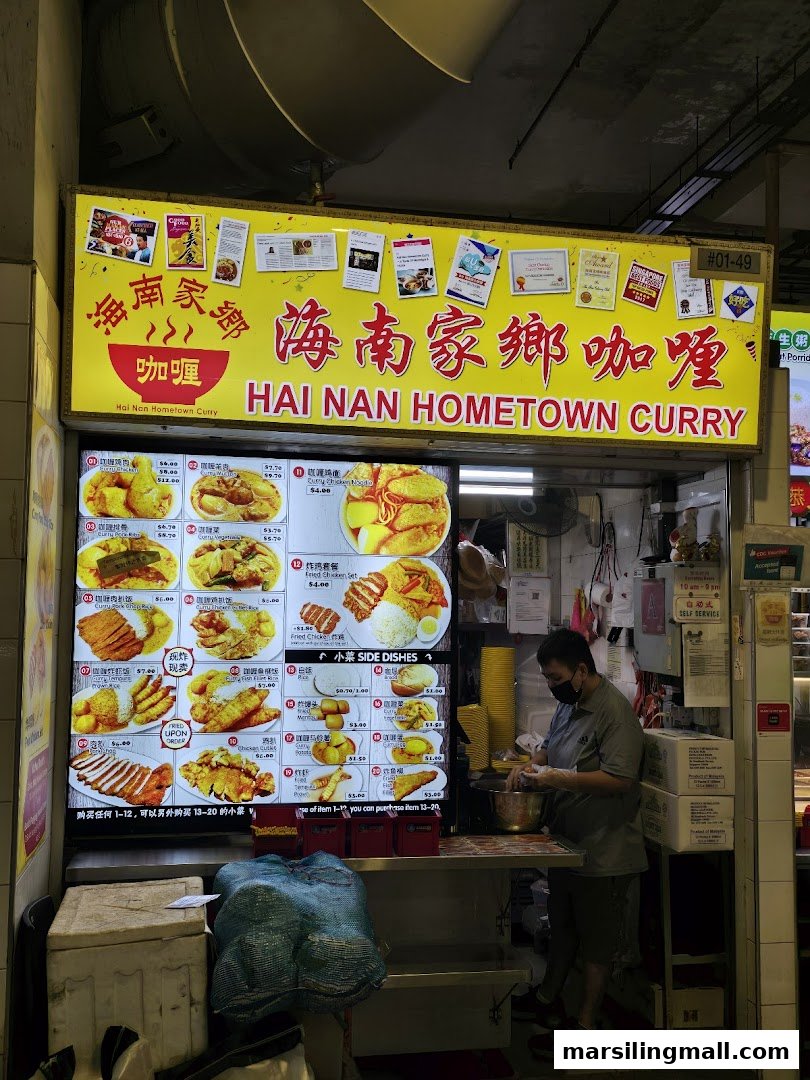 A vibrant food stall named Hai Nan Hometown Curry, featuring a large illuminated menu board and a staff member.