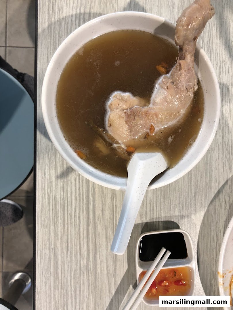 A bowl of herbal chicken soup with a chicken leg, spoon, and dipping sauces on a table.