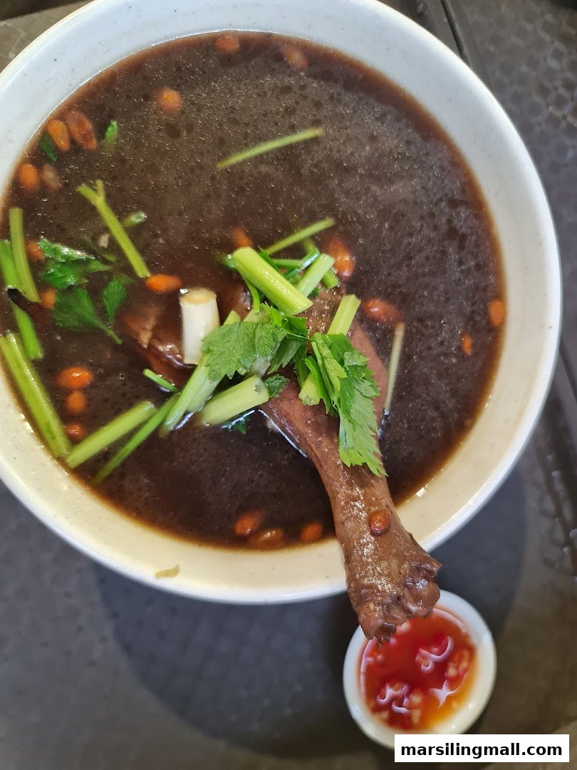 A close-up of a rich herbal duck soup with goji berries, celery, and a side of chili sauce.