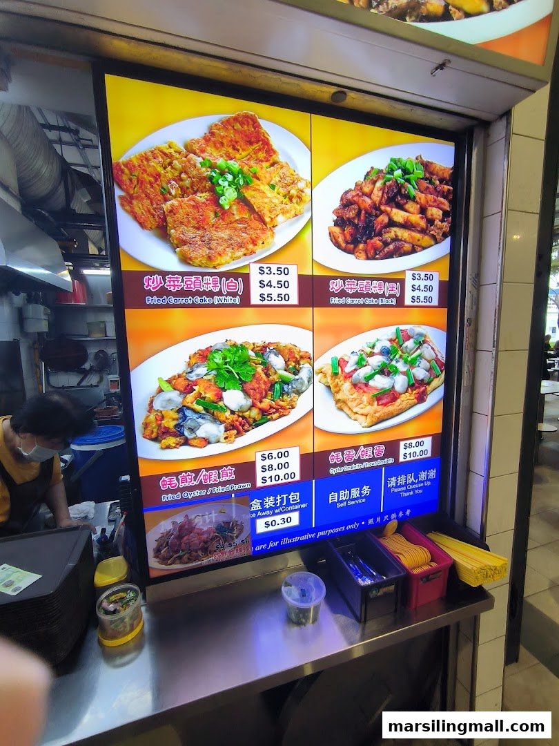 A brightly lit digital menu board displaying various fried carrot cake and oyster dishes with prices at a hawker stall.