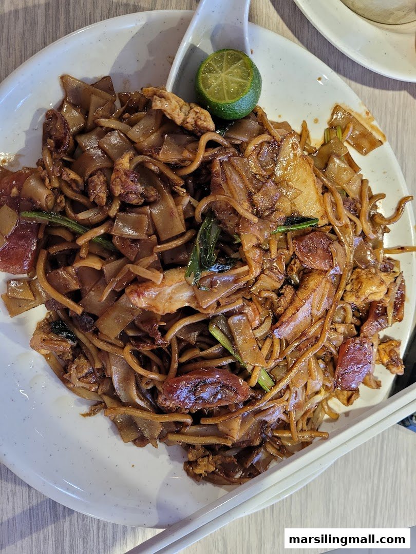 A close-up of a plate of dark, savory fried noodles with egg, meat, and a lime wedge.