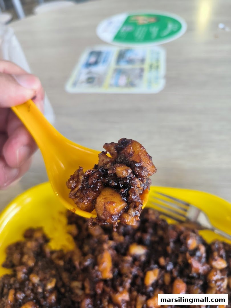 A close-up of dark fried carrot cake on a yellow spoon, with a plate of food in the background.