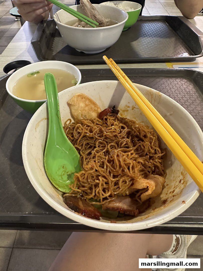 A close-up of delicious dry noodles with char siu and a side soup, with bak kut teh in the background.