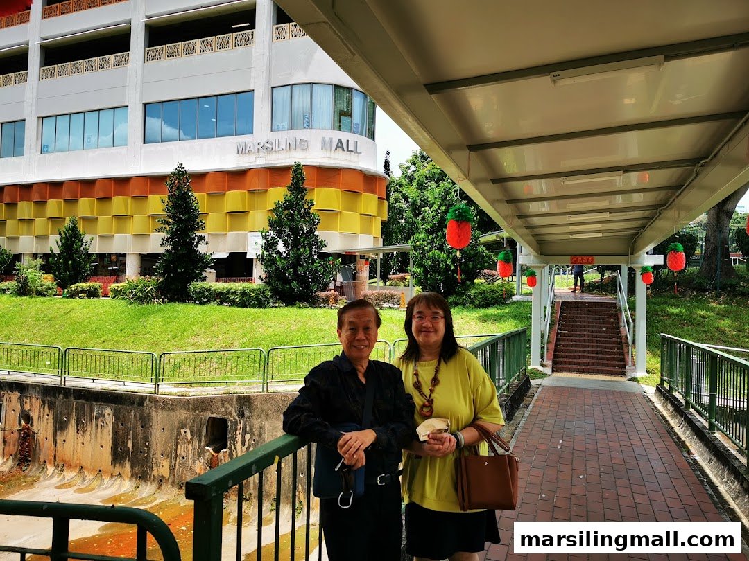Two people stand on a covered walkway with Marsiling Mall and lush greenery in the background.
