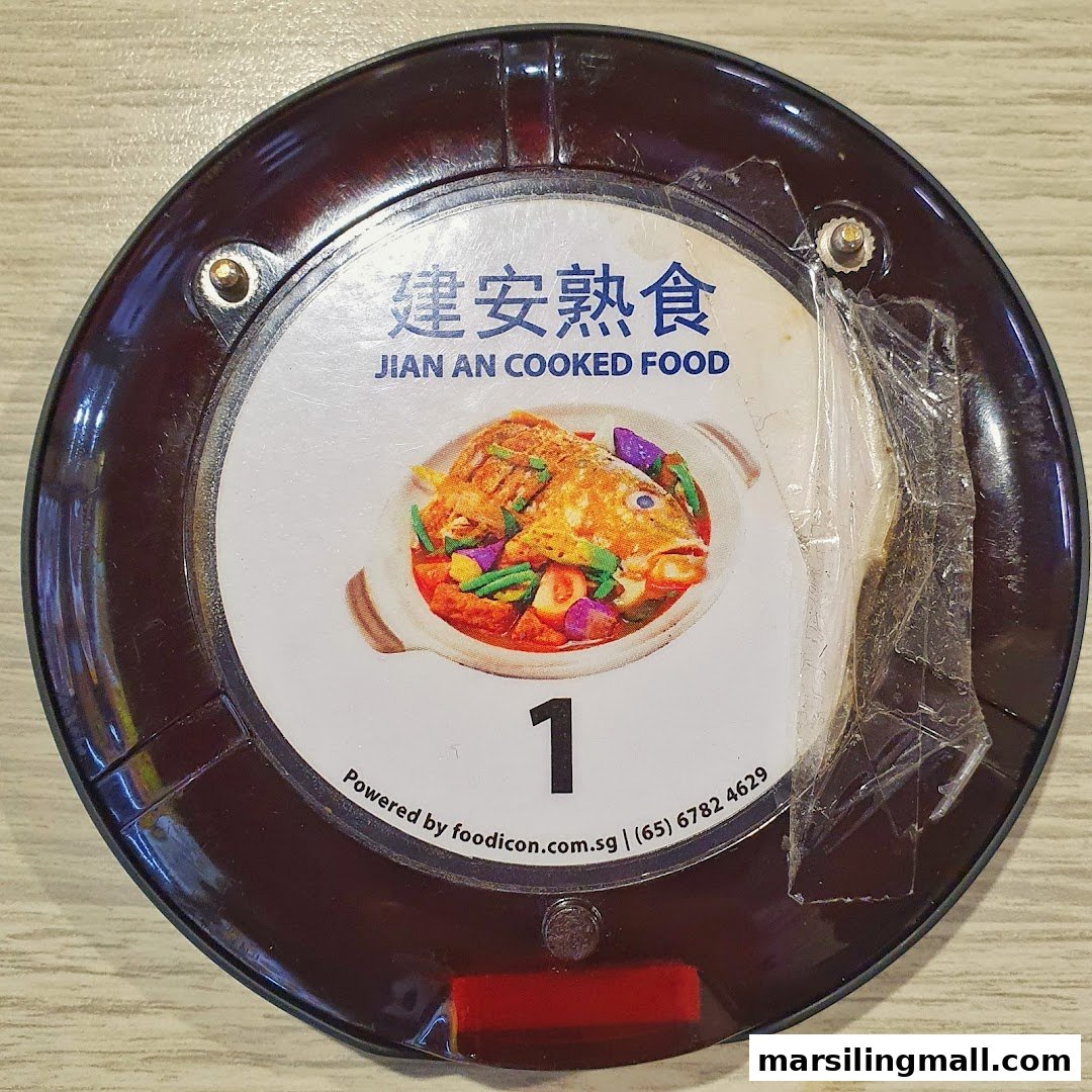 A close-up of a restaurant pager from Jian An Cooked Food, featuring a vibrant fish dish.