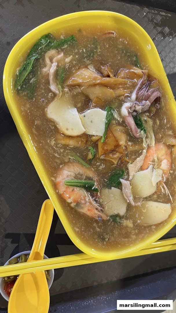 A vibrant yellow bowl filled with a hearty seafood noodle dish, featuring prawns, squid, fish cake, and greens in a rich gravy.