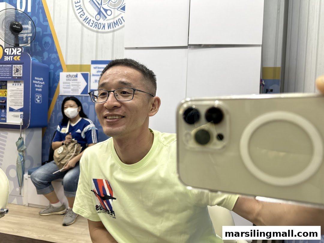 A man takes a selfie inside kcuts, with a woman waiting in the background.