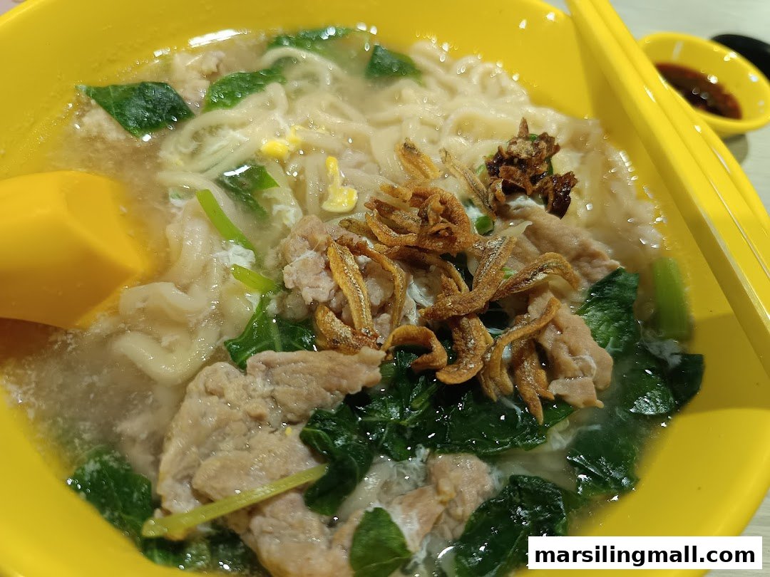 A vibrant yellow bowl of savory noodles with pork slices, greens, and crispy anchovies.