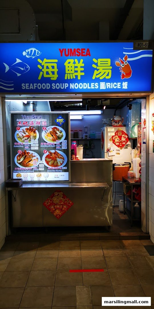 A vibrant hawker stall named YumSea, specializing in seafood soup noodles, with a visible menu board.