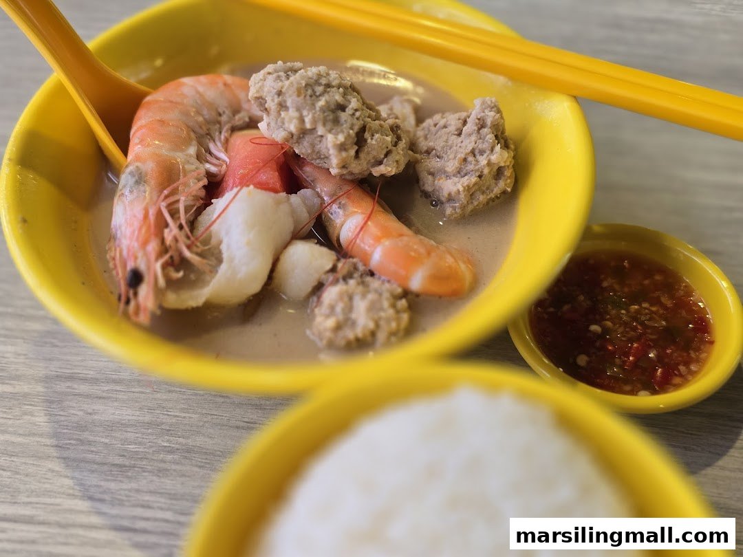 A vibrant yellow bowl of seafood and meatball soup with rice and chili sauce.