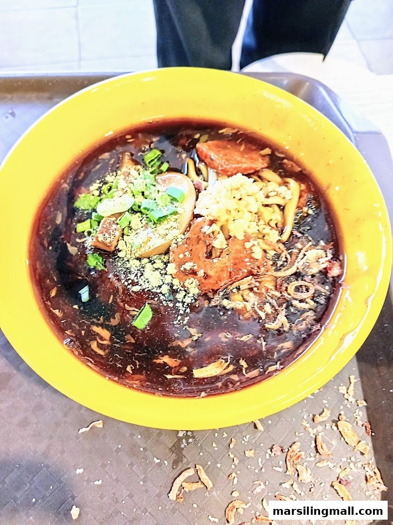 A vibrant yellow bowl filled with dark sauced noodles and various toppings, including fried shallots and greens.
