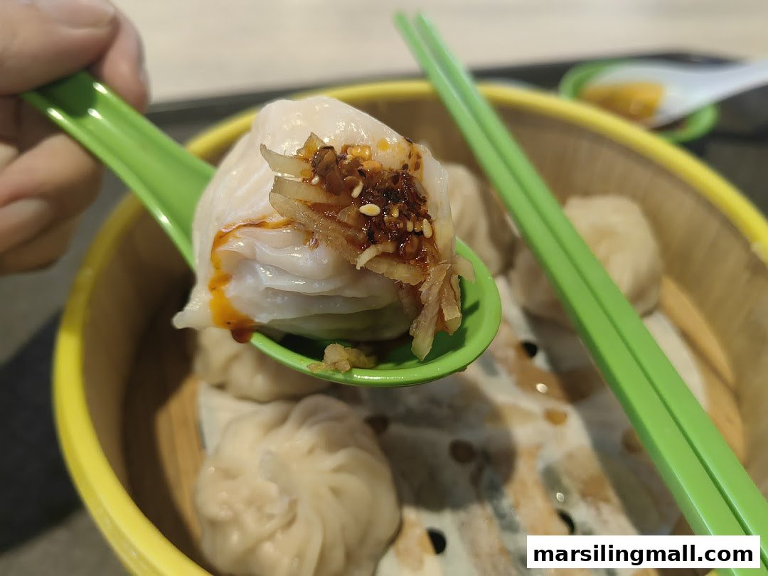 A hand holds a steamed dumpling with chili oil and ginger on a spoon, from a bamboo steamer.