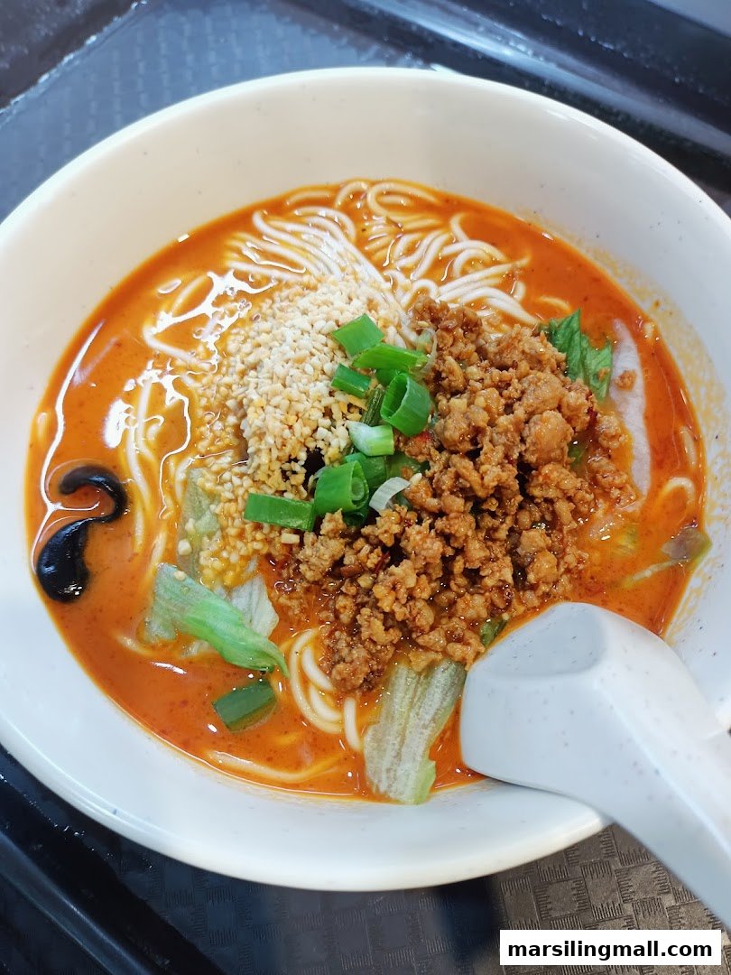 A close-up of a bowl of spicy noodles with minced meat, peanuts, and greens.