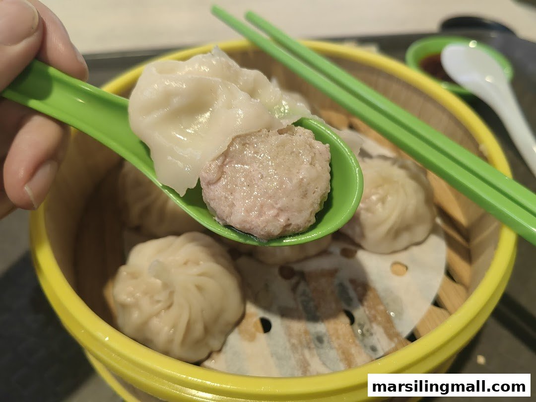 A hand lifts a freshly steamed xiao long bao from a bamboo basket with a green spoon.