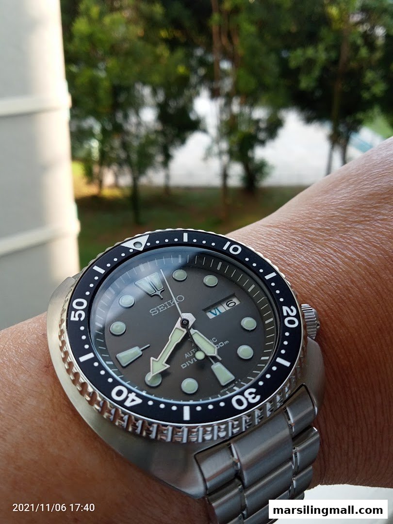 A close-up of a Seiko watch on a wrist, with a blurred outdoor background.