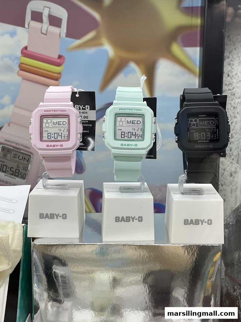 Three colorful Baby-G watches, pink, mint, and black, displayed on white stands.