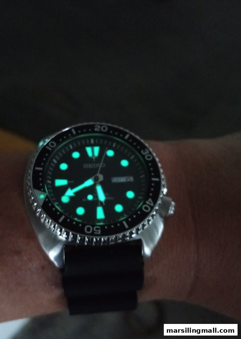 A close-up of a luminous Seiko watch on a wrist in low light.