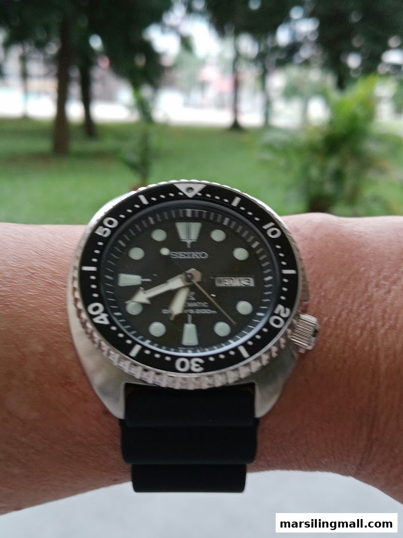 A close-up of a Seiko dive watch on a wrist against a blurred green outdoor background.