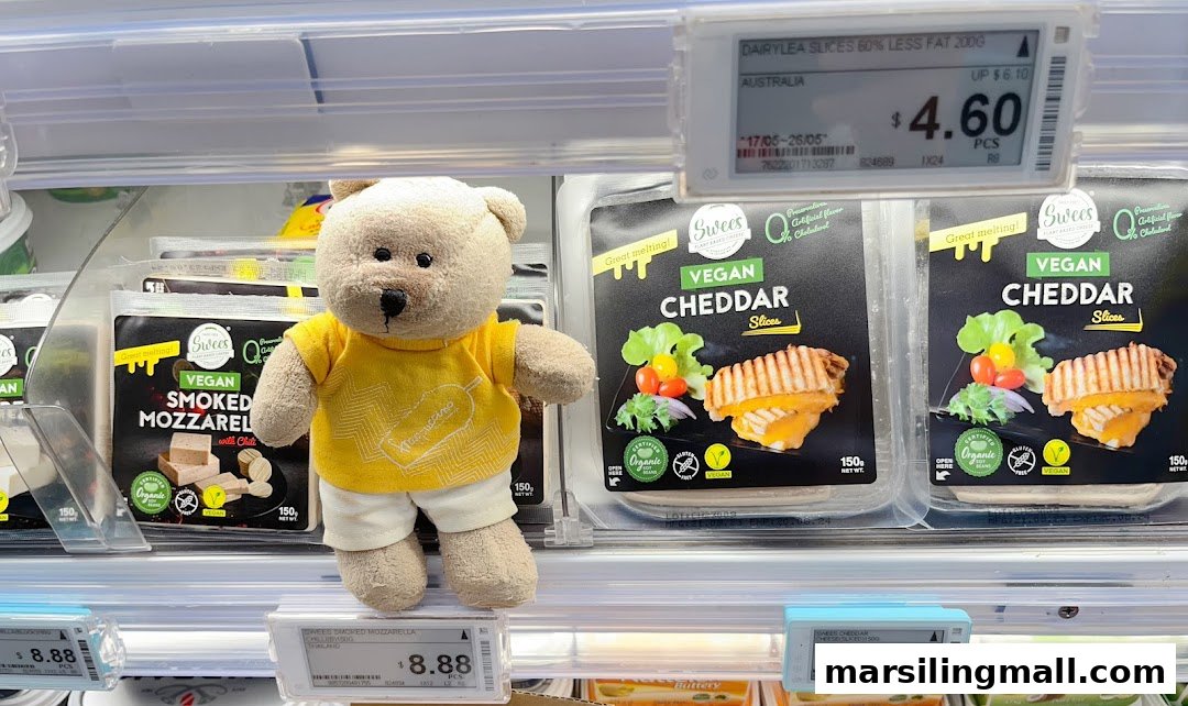 A teddy bear stands among vegan cheese products and price tags in a supermarket aisle.