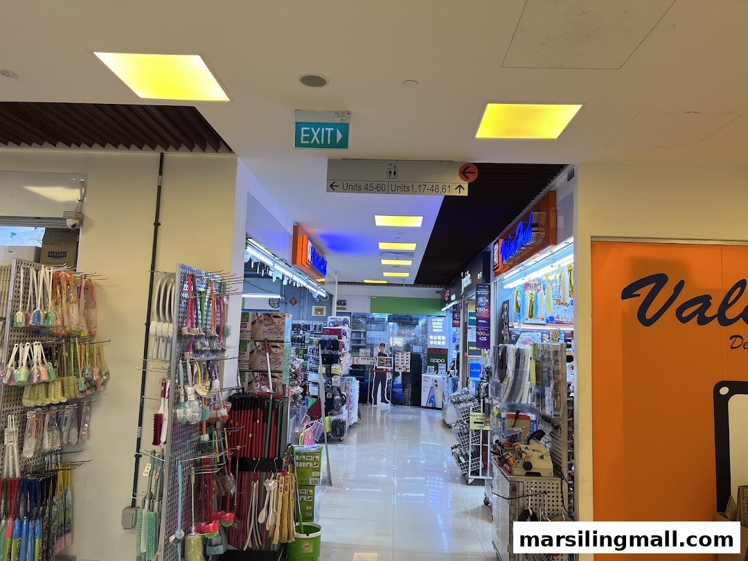 An interior view of a shopping mall aisle featuring various retail shops and household items on display.