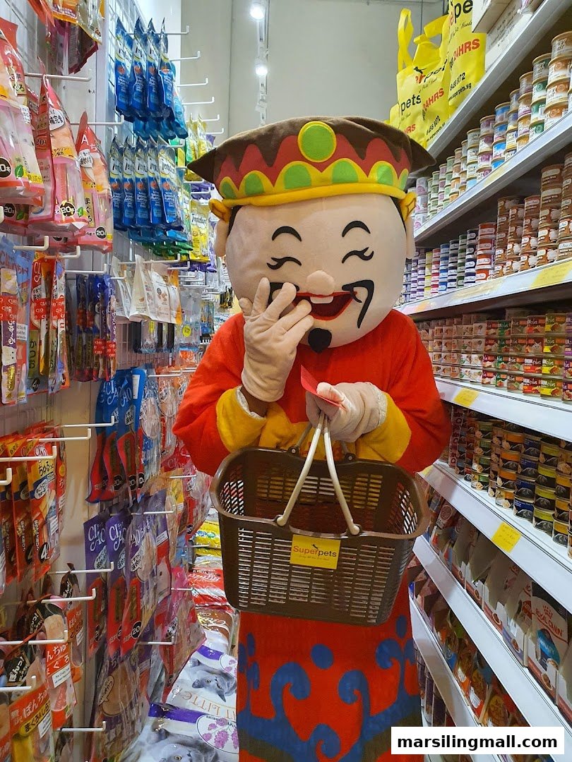 A cheerful mascot in traditional attire holds a shopping basket amidst pet food and supplies at Superpets.