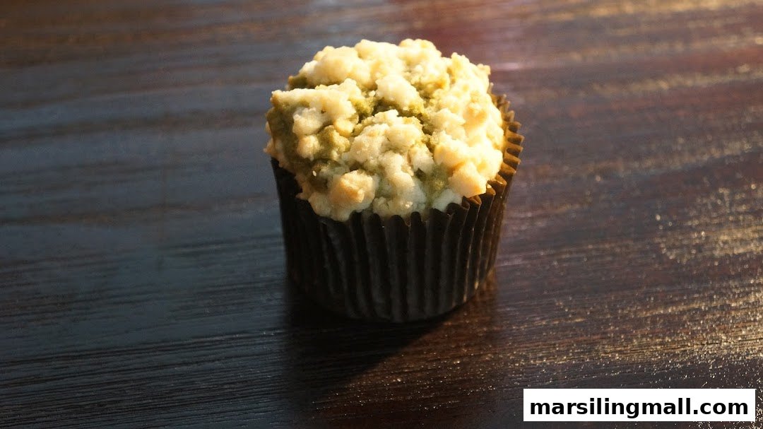 A delicious-looking cupcake with a crumbly, possibly pistachio or matcha, topping sits on a dark wooden surface.
