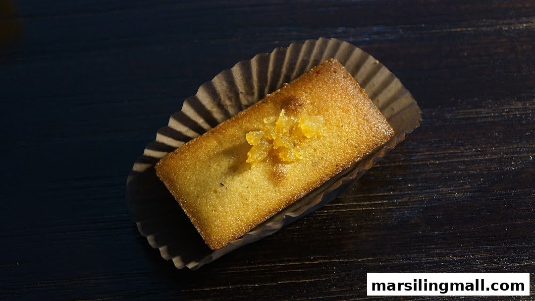 A golden-brown financier or small cake with candied fruit topping in a paper liner on a dark wooden surface.