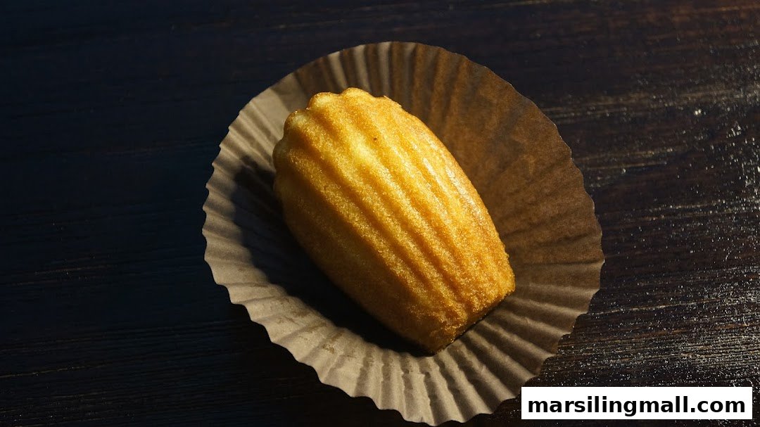 A golden-baked Madeleine cookie rests in a fluted paper cup on a dark wooden surface.