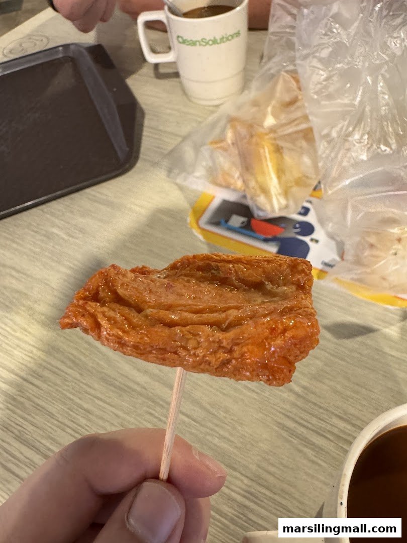 A hand holding a fried fish cake on a stick, with drinks and other items in the background.