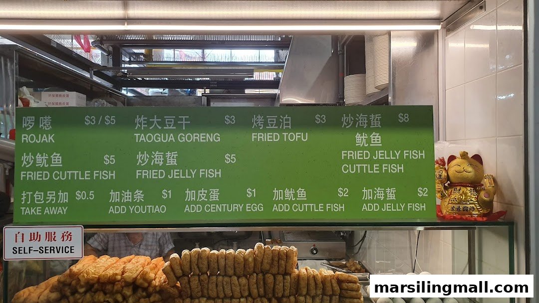 A green menu board for Woodlands Rojak displaying various rojak and fried items with prices, above a food counter.