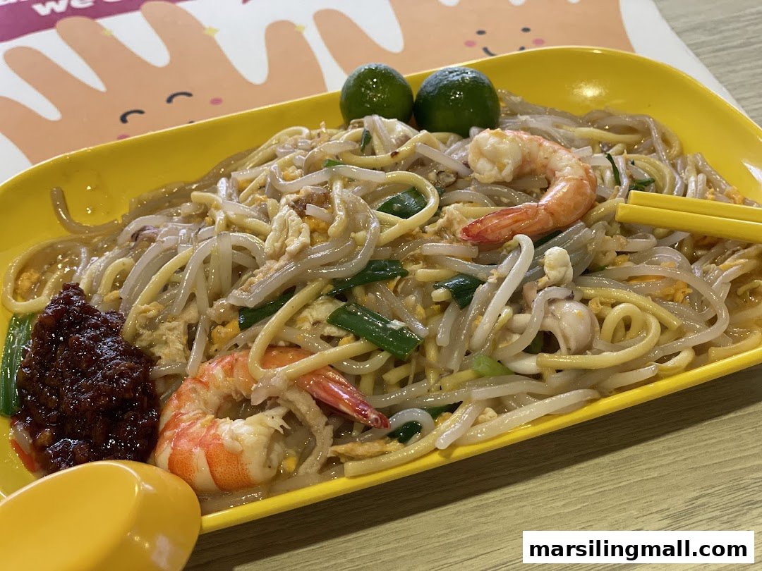 A vibrant plate of Hokkien Mee with prawns, squid, and a side of sambal chili, garnished with calamansi limes.