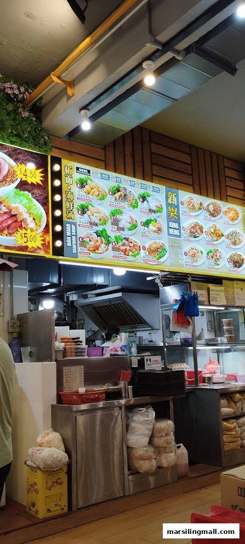 A bustling food stall counter featuring a large menu board displaying various fried noodle and soup dishes.